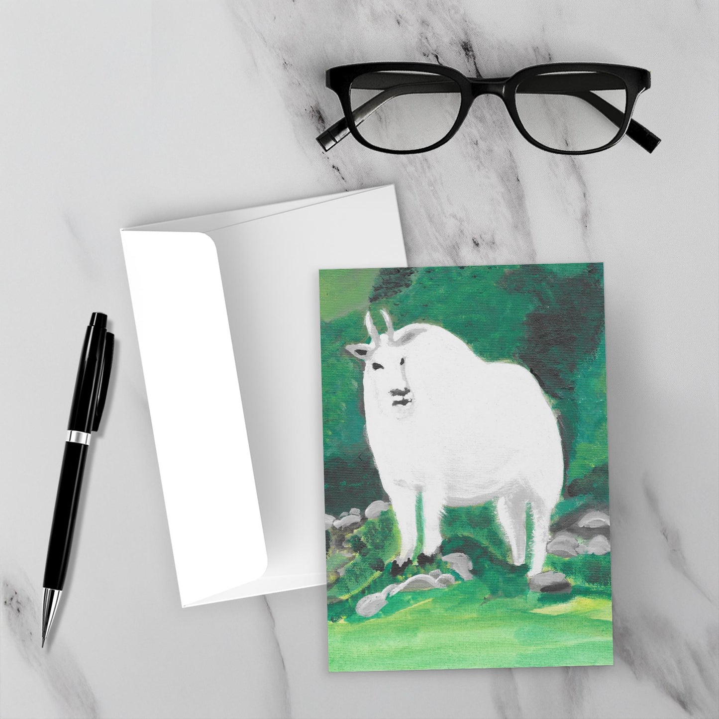 Mockup display showcasing the front of the greeting card Mt. Rushmore Mountain Goat lying on a gray marble surface along side a white envelope, a pair of black reading glasses and a black pen.