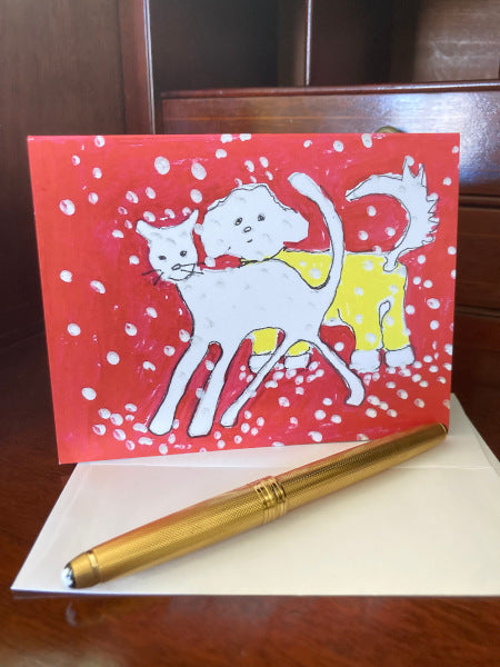 Photograph of the winter scene greeting card of a white cat and a white dog in a yellow with snowflakes falling against a vivid red backdrop. The card is standing upright on a brown desk with a white envelope and a gold pen in the foreground.