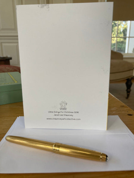 Photograph of the back of the holiday greeting card titled Little Doings For Christmas which shows the card back with logo and title standing on the desktop with envelope partially showing and a gold pen.