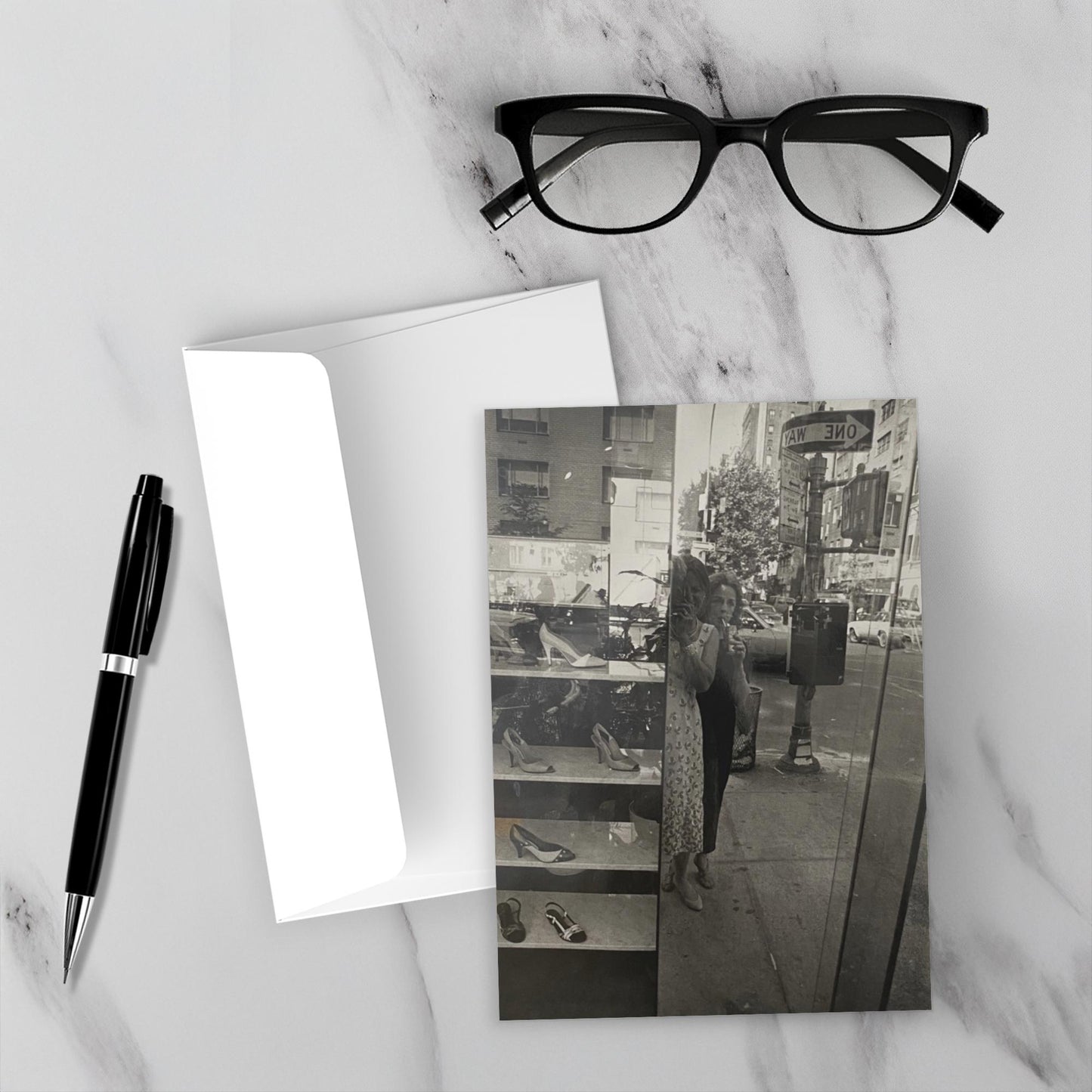 Mockup of Madison Avenue Shoe Shopping on a marble backdrop. The envelope, a pen and glasses are also displayed.
