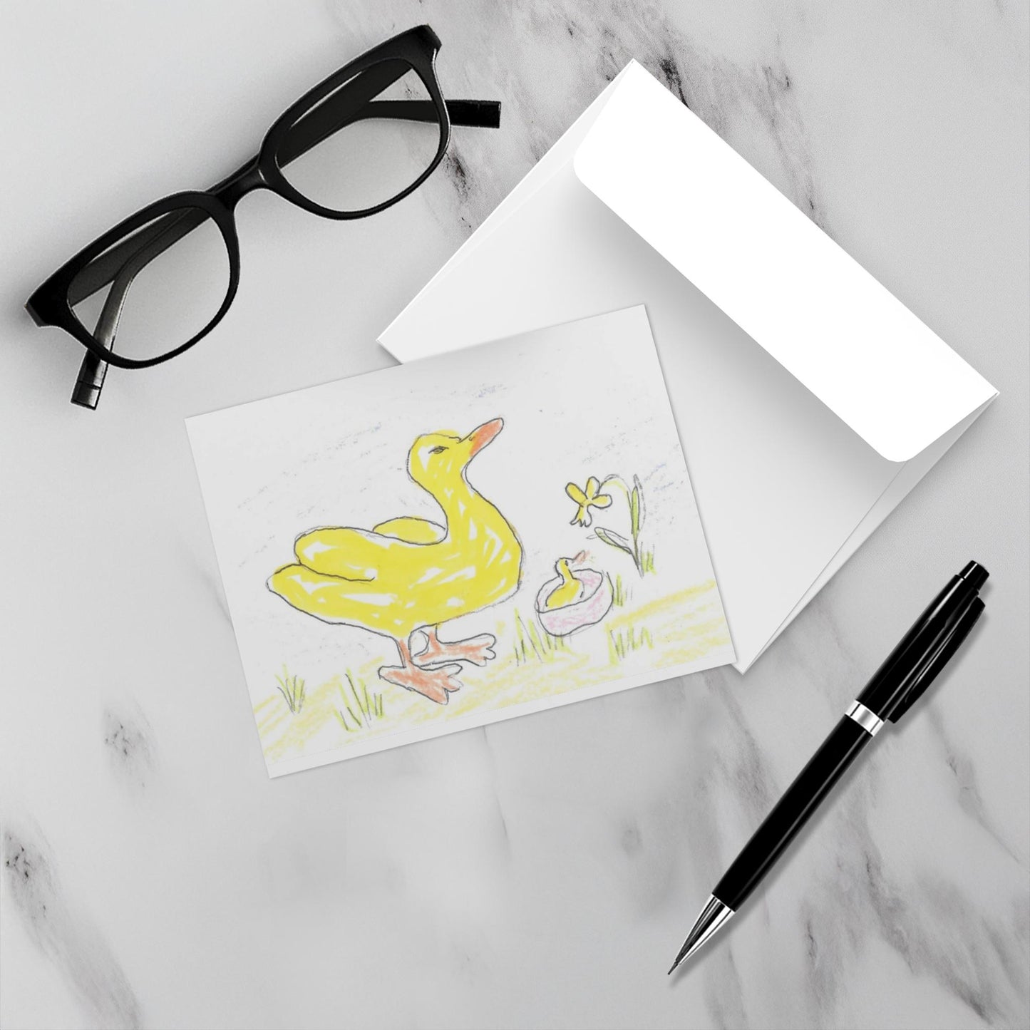 Mockup display showcasing the front of the greeting card Two Yellow Ducks One Yellow Flower lying on a gray marble surface along side a white envelope, a pair of black reading glasses and a black pen.