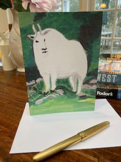 The front of the Card Mt. Rushmore Mountain Goat displayed on a brown desk with a white envelope and gold pen in the foreground and travel books barely visible in the background.