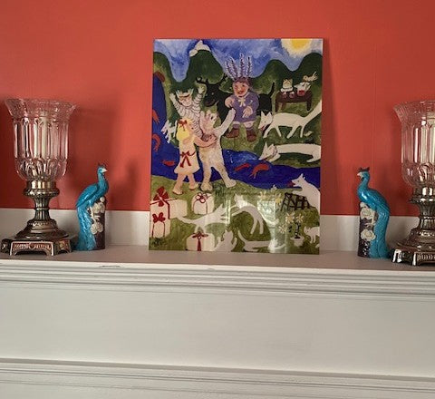 Mockup of the acrylic art print titled Party Time is photographed here propped on a mantle with two bird statuettes and two hurricane lamps. The artwork shows a fanciful outdoor birthday party picnic with wrapped presents in the foreground, human and animal guests arriving and being greeted by the birthday boy. River and mountains frame the scene.