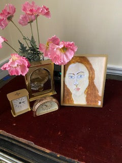 Mockup of the art print Portrait Girl in an easel-backed gold frame on a red velvet topped writing desk next to three decorative gold clocks and a vase of pink poppies.
