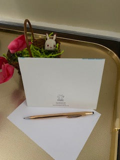 Back of the card Prissy Egg Walk displayed on a gold colored tray with a little Easter Basket that holds a little Easter bunny ornament and some red flowers in the background and a white envelope and a gold pen in the foreground.