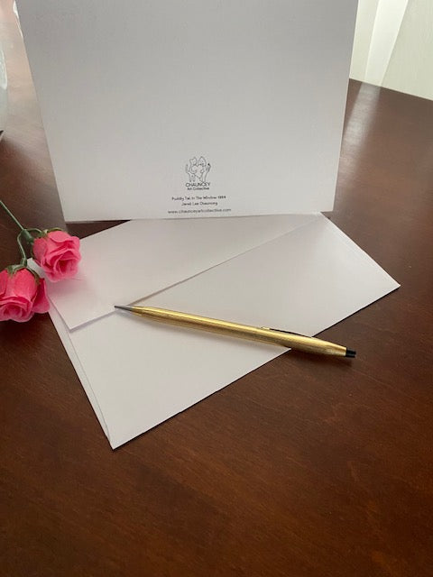 Photograph display of the back of a black and white greeting card titled Puddy Tat In The Window arranged on a brown desktop with a white envelope, gold pen, and two pink rosebuds.
