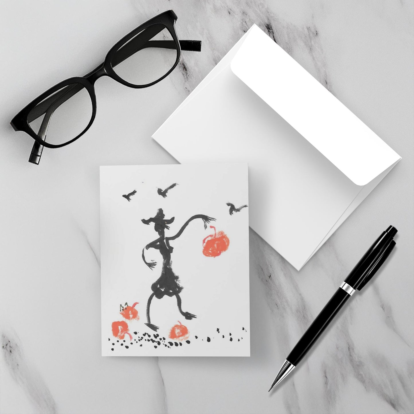 Mockup of the halloween card Pumpkin Drop which shows the front of the card, the back of the card and a black pen and a pair of black glasses ton a grey marble surface.