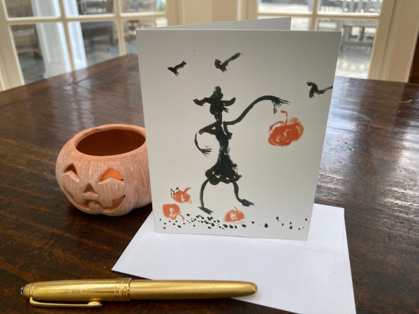 photograph of the front of the halloween card Pumpkin Drop which is upright on brown tabletop atop it's white envelope with a gold pen in the foreground and a ceramic orange jack-o-lantern to the left.