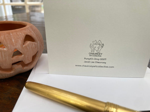 photograph of the back of the greeting card titled Pumpkin Drop. The card is upright on the table top with white envelope and next to an orange ceramic jack-o-lantern. A gold pen is partially in view.