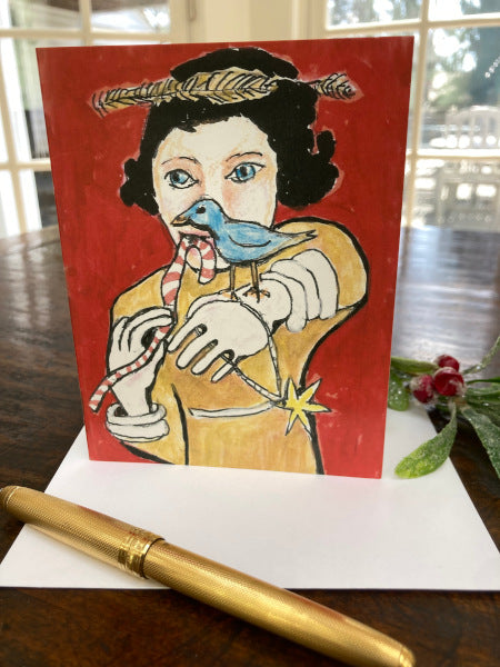 photo of the holiday greeting card titled Red Holiday Girl With Bird which is standing upright on the desktop with white envelope, gold pen and sprig of frosted holly.