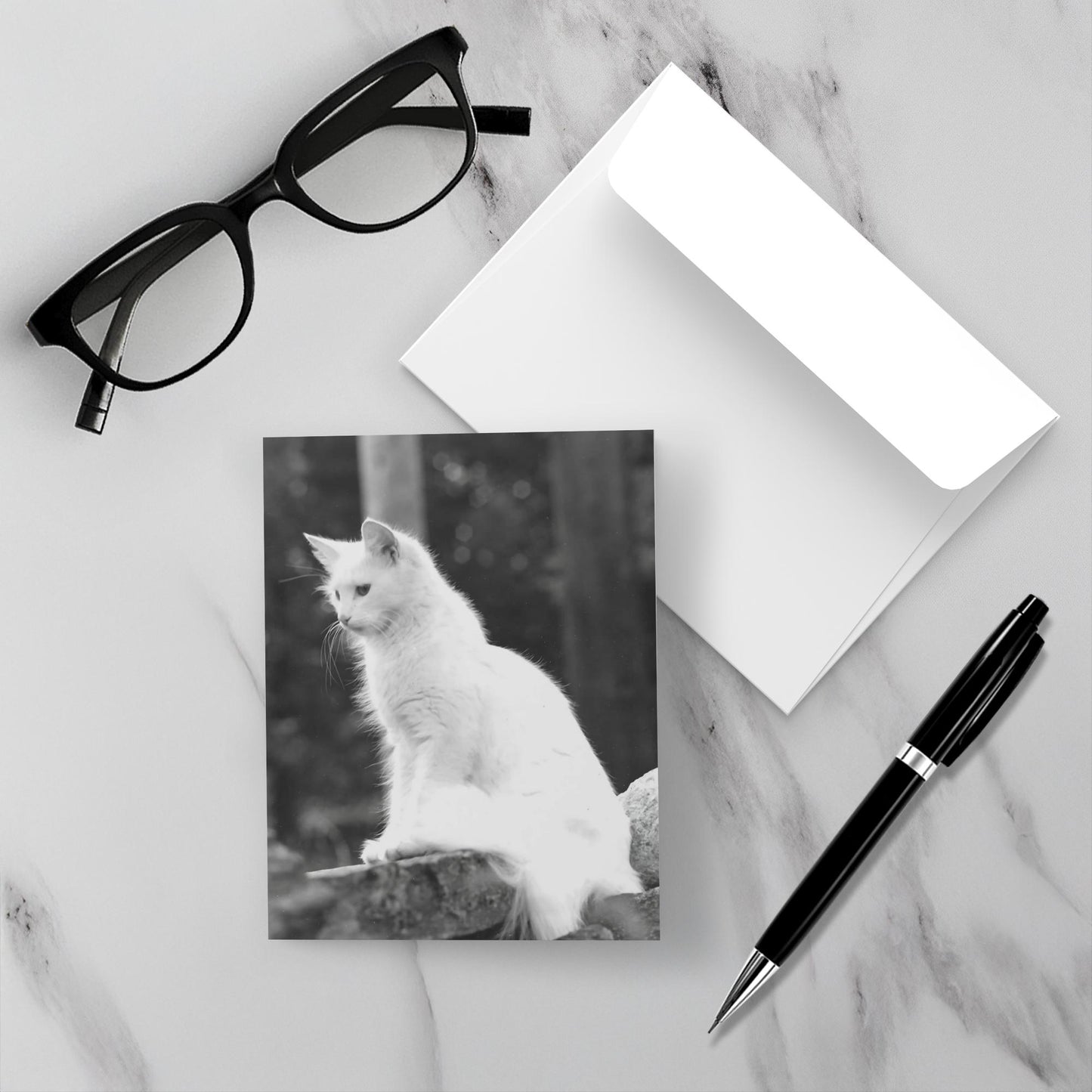 The Roxanne card is face up on a white marble backdrop along with the envelope a pen and reading glasses.