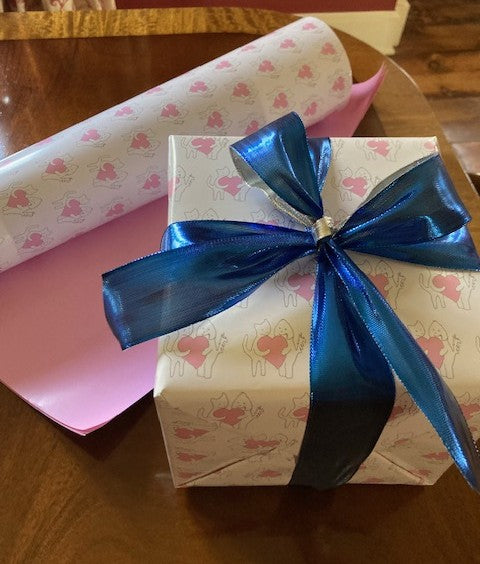 A present is elegantly wrapped in Sharing A Big Heart wrapping paper and tied with a blue ribbon. An extra piece of the sheet paper is in the background showing the front and pink reverse side of the gift wrap.