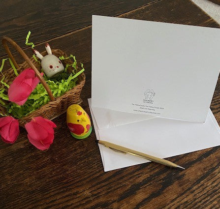 The back of Two Yellow Ducks One Yellow Flower greeting card for spring is displayed on a brown surface with a white envelope, gold pen and a small easter basket filled with green fake grass, wooden easter ornaments in the shape of a bunny and a chick and three red flowers.