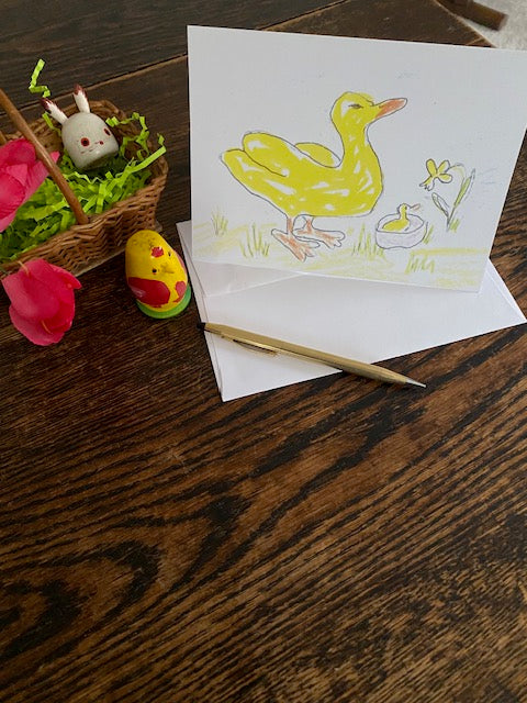 The front of Two Yellow Ducks One Yellow Flower greeting card for spring is displayed on a brown surface with a white envelope, gold pen and a small easter basket with wooden easter ornaments of a bunny and a chick green fake grass and two red flowers.