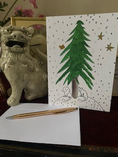 Mockup of Under The Tree Christmas card on a desk with a gold pen and the envelope sitting next to a white foo dog.