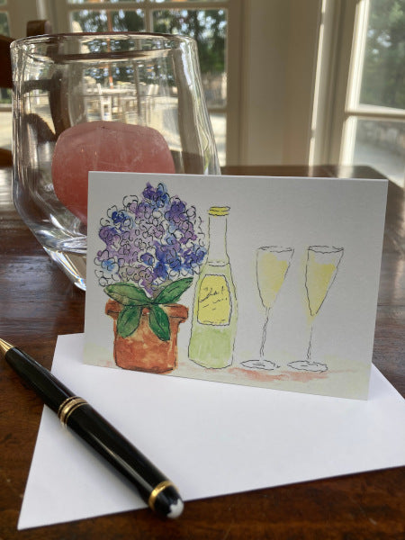 The front of Wine Time notecard is with its envelope sitting on a wooden desk with a fountain pen and a glass hurricane lamp with a rose-colored candle.