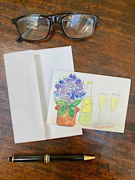 The front of Wine Time notecard sitting atop a wooden table with its envelope. Dark brown glasses and a brown fountain pen are also shown.