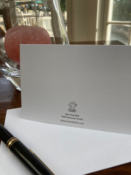 A mock up of the back of the notecard Wine Time with its envelope on a wooden desk with a hurricane lamp and rose-colored candle in the background. Chauncey Art Collective's logo is also featured.