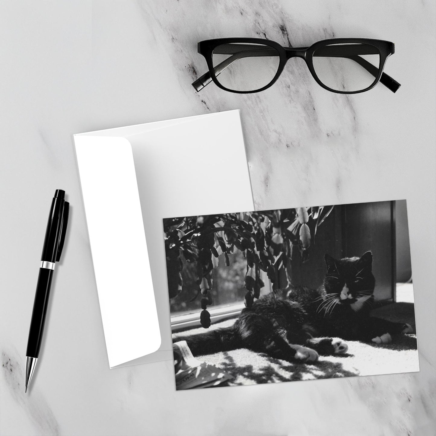 Mockup display showcasing the front of the greeting card Puddy Tat In The Window lying on a gray marble surface along side a white envelope, a pair of black reading glasses and a black pen.