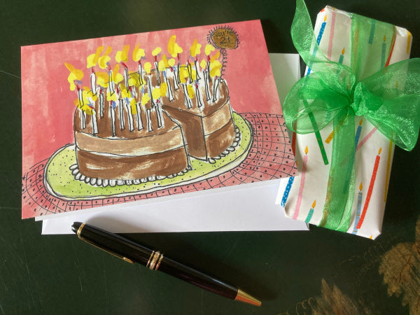 Mock up of You're 21! birthday card. Close-up of a decadent chocolate cake, its surface dotted with burning candles. A little sign propped against the frosting proudly announces "You're 21". A wrapped present and fountain pen are also shown.