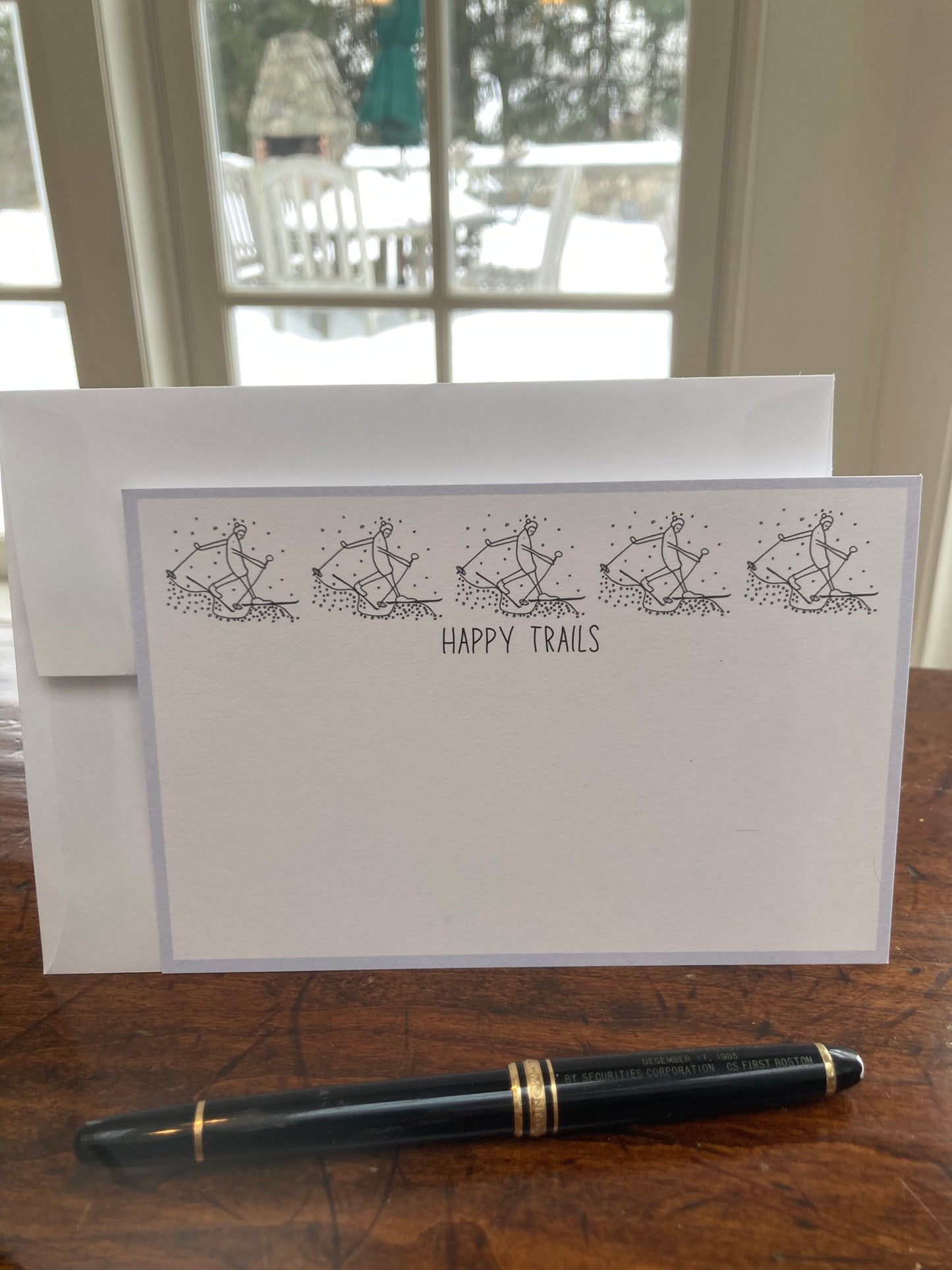 Flat note card with 'Happy Trails' text and illustrations on a wooden surface with a pen.