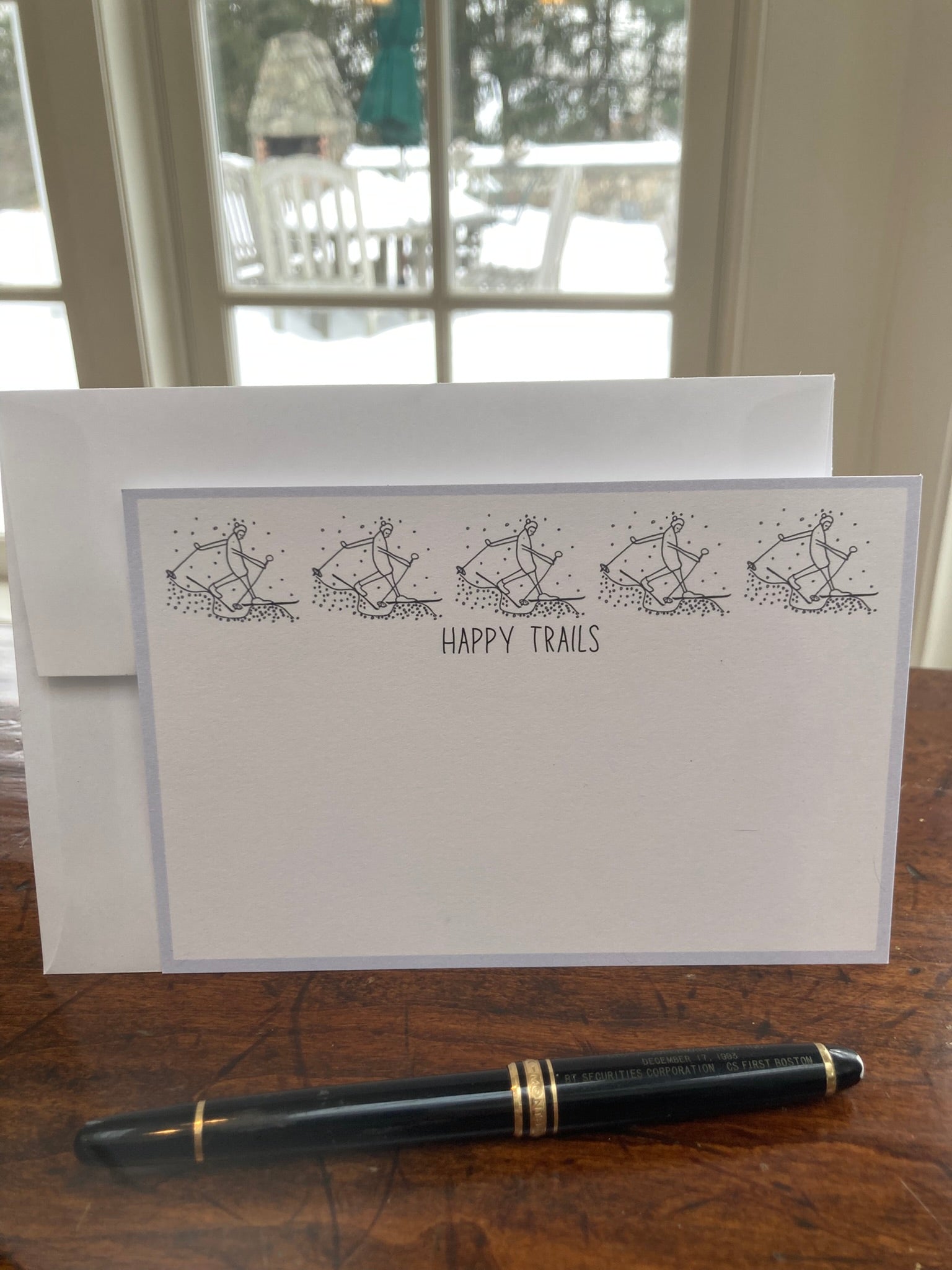 Flat note card with 'Happy Trails' text and illustrations on a wooden surface with a pen.