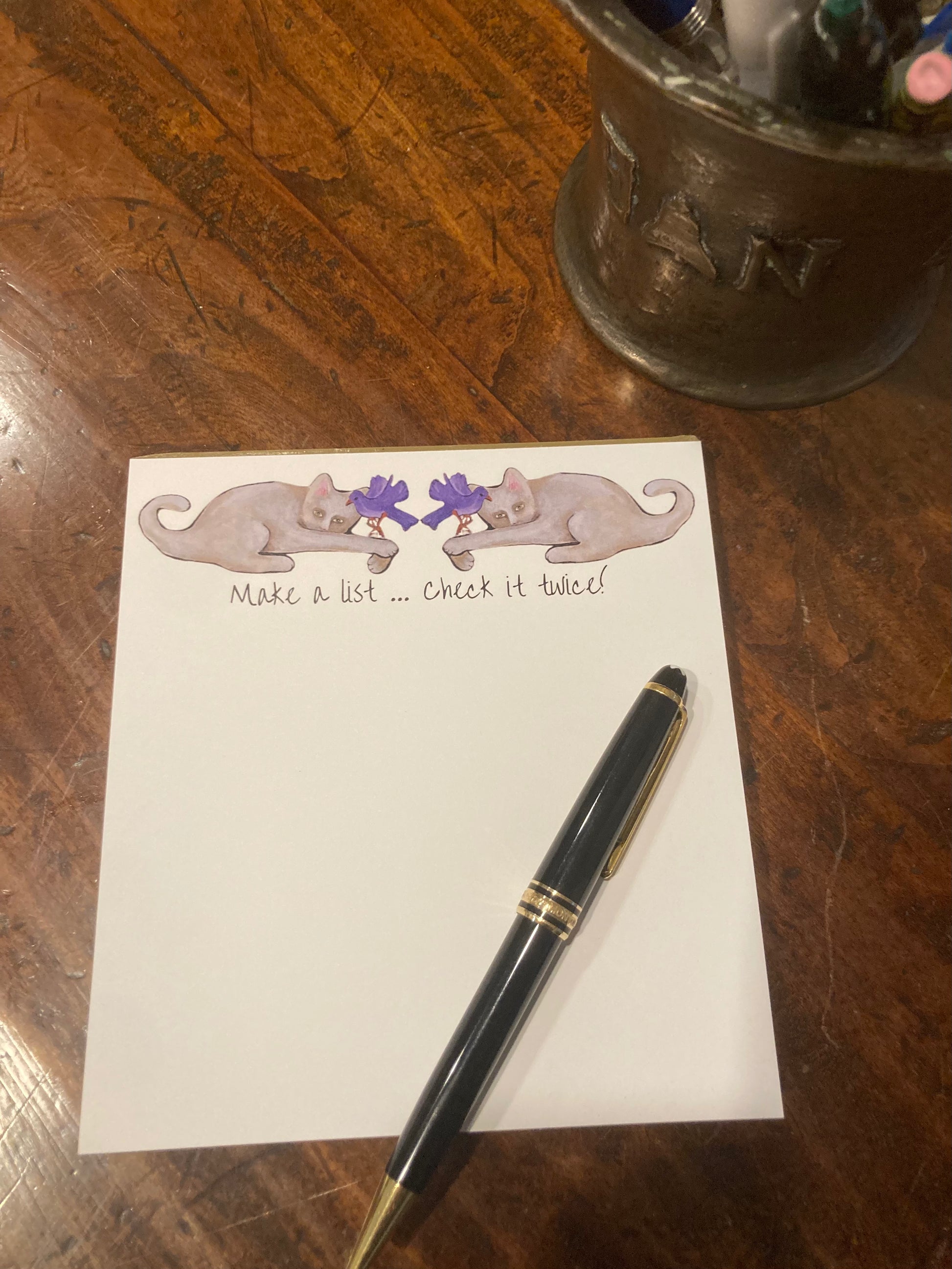 White notepad with illustration of two cats holding purple birds and a pen on a wooden surface.