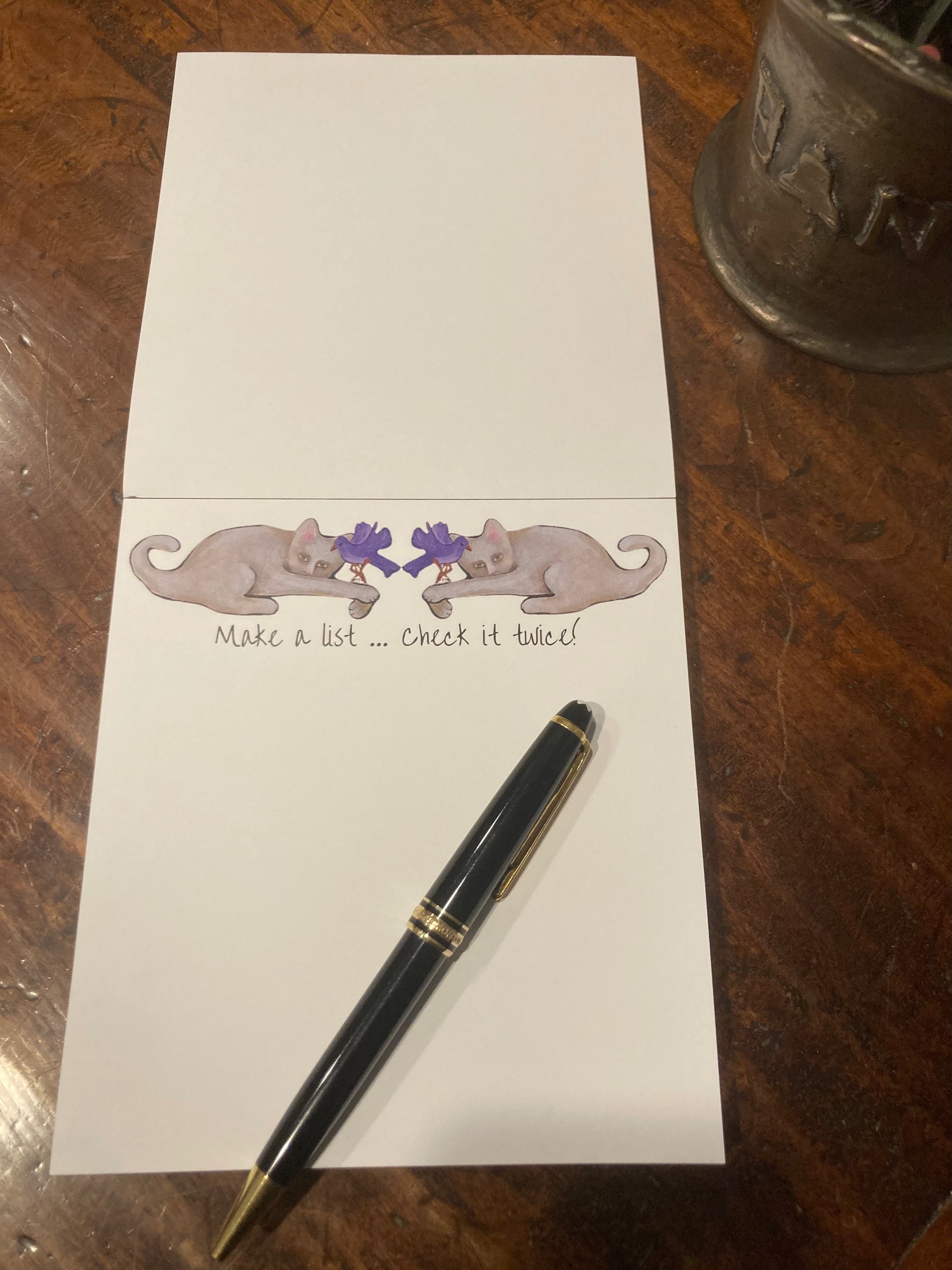 Notepad with decorative cat and purple bird design and pen on a wooden surface.