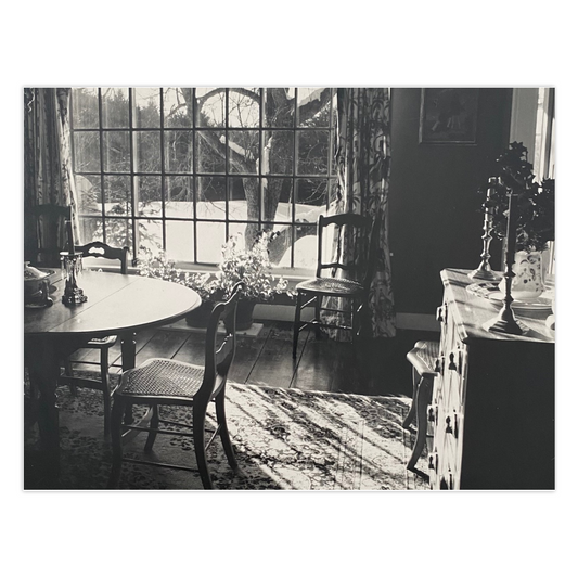 Front of the photo greeting card titled Vermont Light showing an interior of a country home dining room with partial view of wooden table, cane seat chairs, oriental rug on the wide planked floor,  crystal candlesticks on the table, floral drapes, and a side bureau with pitcher, plate and brass candlesticks.  There is a large paned glass window through which a snowy winter landscape is visible, most notably a large trunk and limbs of an apple tree.  The title of this winter scene card is Vermont Light.