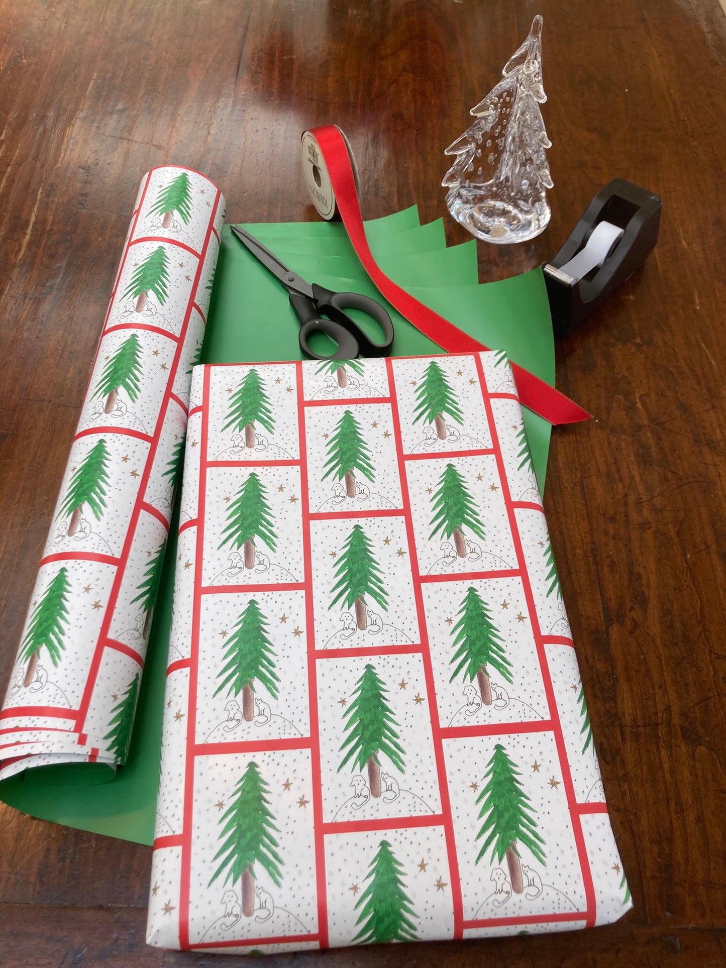 Mockup of the Under The Tree Wrapping paper showing a shirt box wrapped in gift paper lying atop the unfurled sheets of paper showing the front and the plain green backside.  A pair of scissors, red gift wrap and a tape dispensoe are in the phot along with a glass Christmas tree statuette.