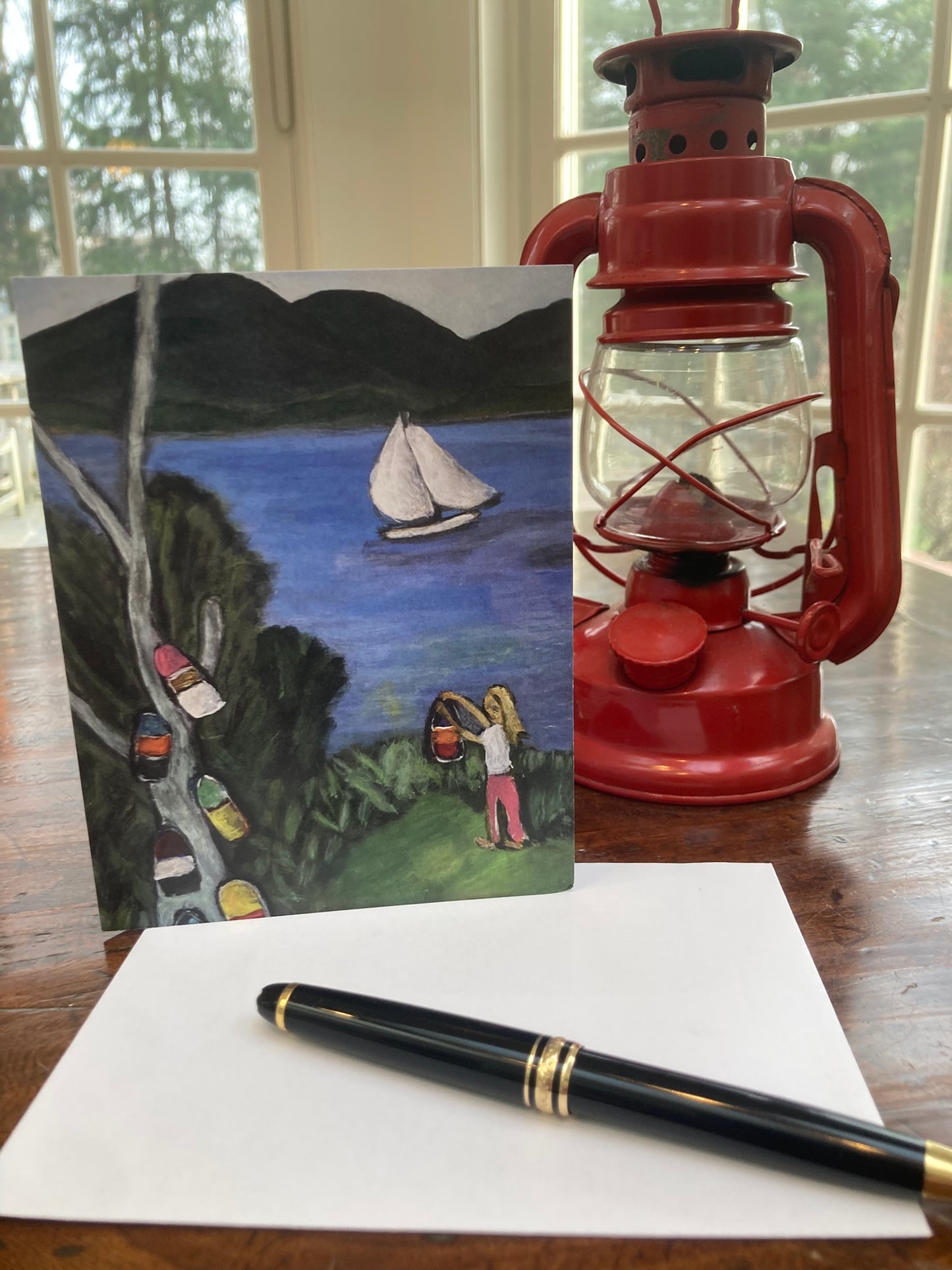 Greeting card of a girl hanging lobster buoys in a tree by the seashore with sailboat and mountains.  The card is on a table next to a red lantern.