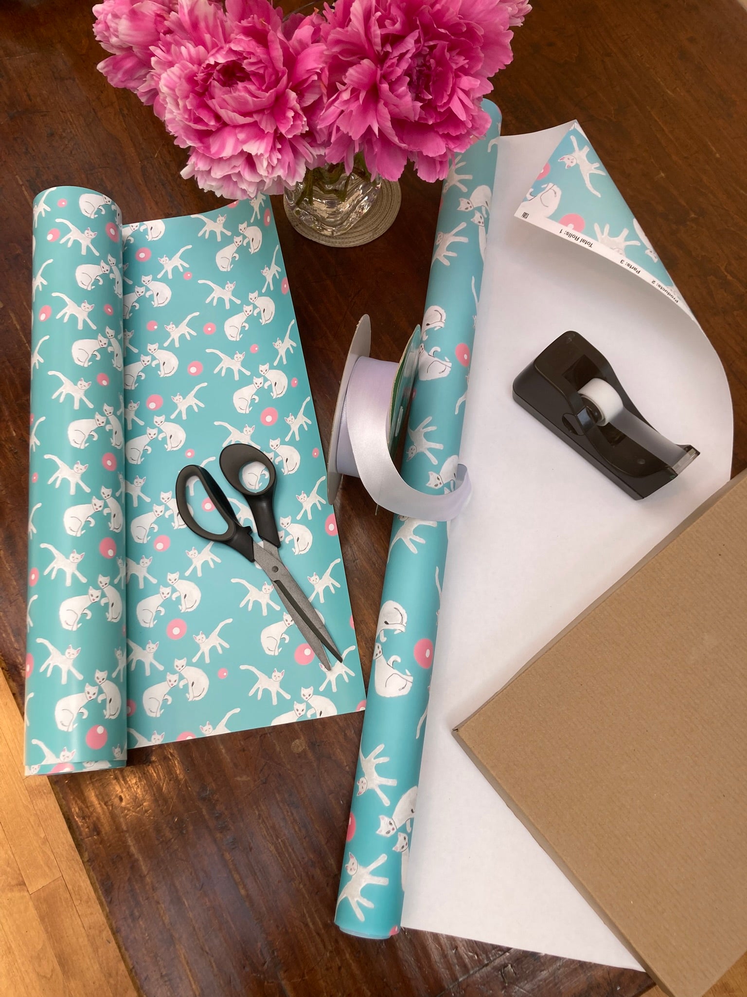 a mockup of the Fancy Felines wrapping paper showing the sheets printed on both sides unfurling and the larger roll of wrapping paper printed on one side with shirt box on top about to be wrapped.  There are scissors, tape dispenser and vase of pink flowers to giver perspective.