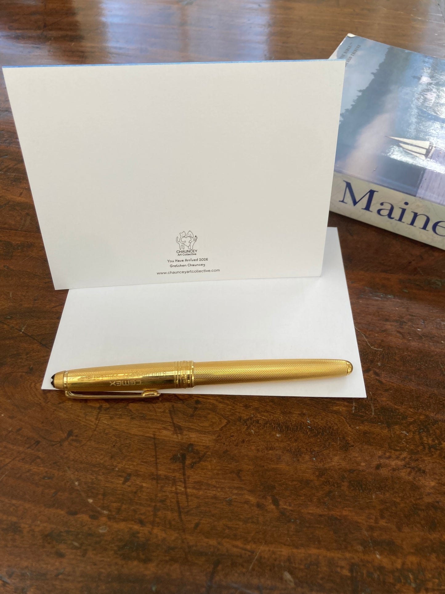 Gold pen on a white card showing the title and the Chauncey Art Collective ogo on a wooden surface.
