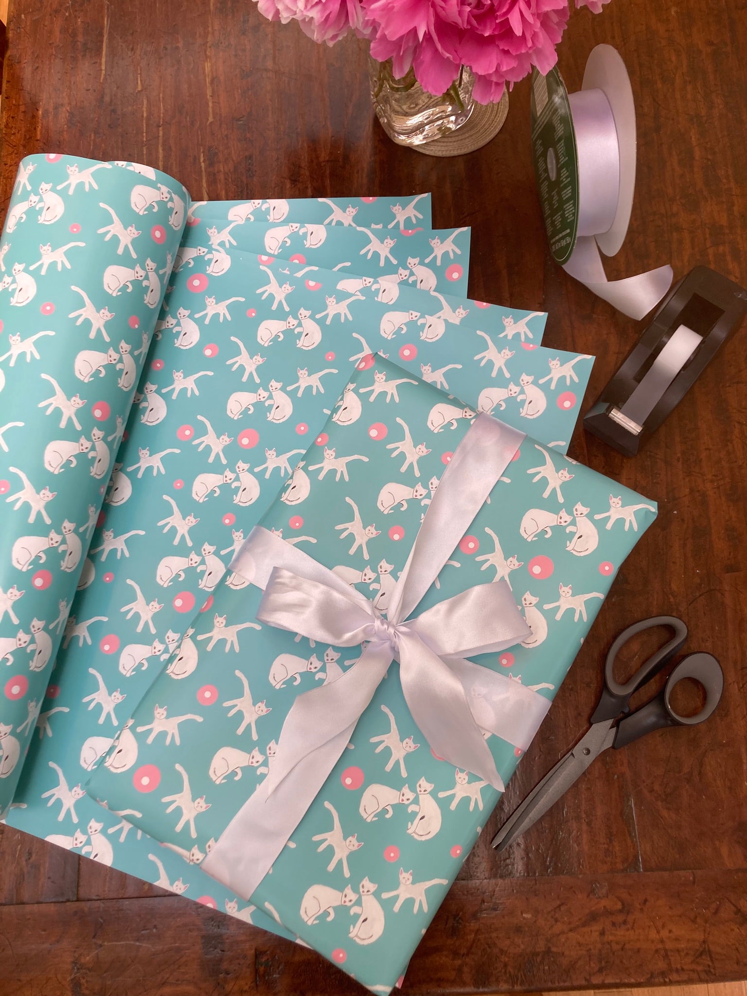 Mockup of the teal blue gift wrap with white cats and pink dots on table top where you see a shirt box wrapped in the paper with a white bow on top of the remaining 4 sheets of wrapping paper along with a tape dispenser and a roll of ribbon and pink flowers partially in view.