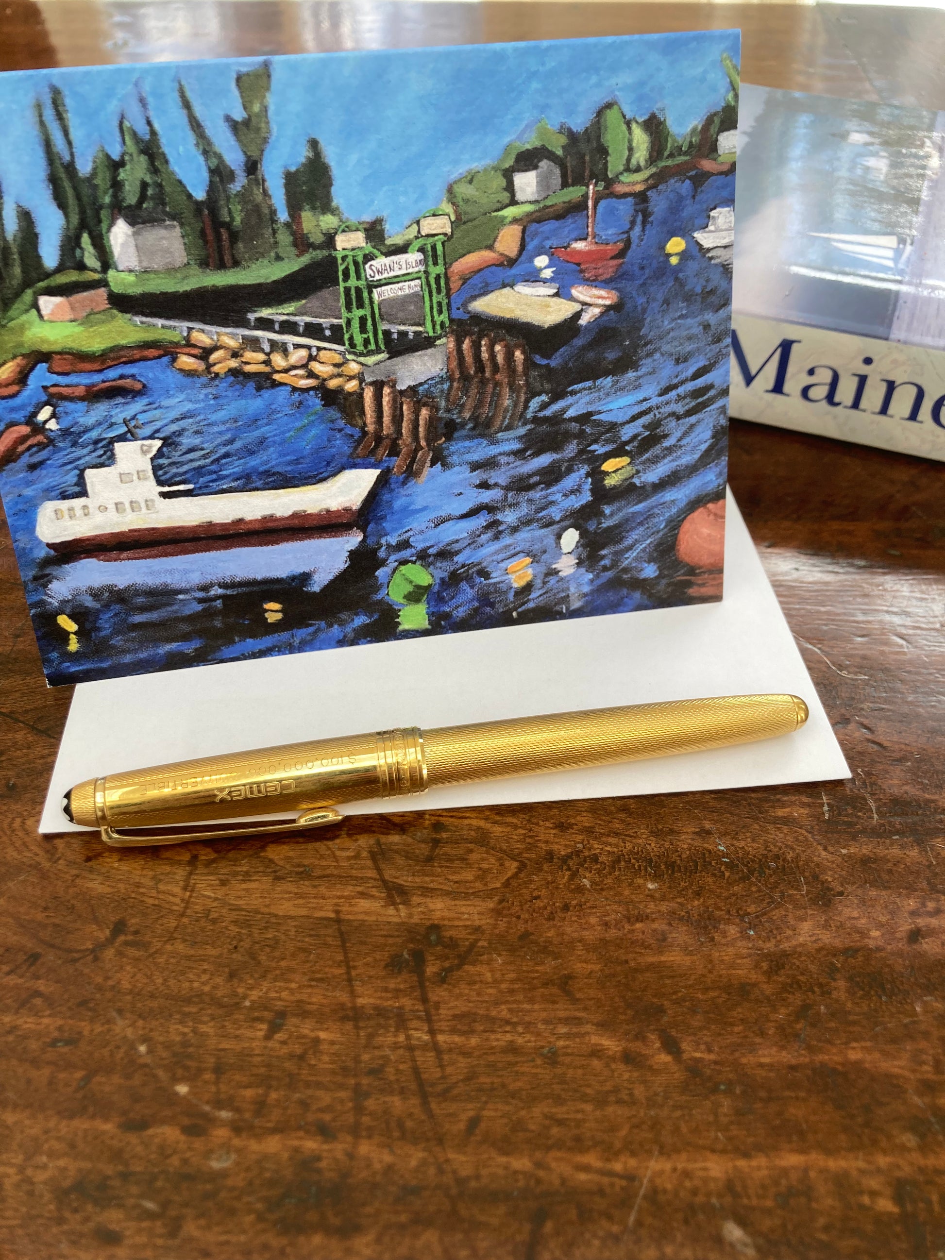 Swan's Island greeting card of a harbor scene by Gretchen Chauncey with a gold pen on a wooden surface.