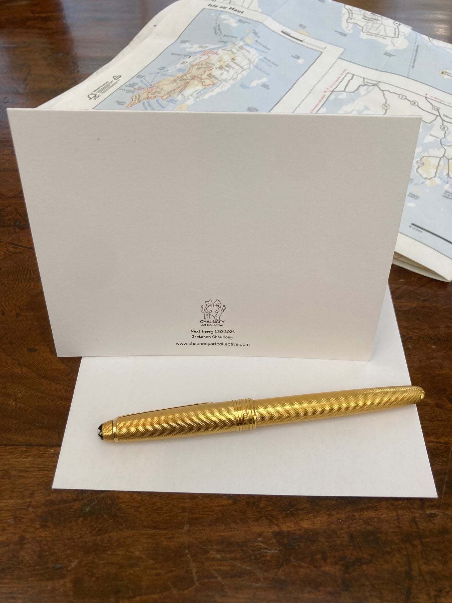 Gold pen with back of Chauncey Art Collective greeting card The Next Ferry 1:30 placed on a wooden surface with maps in the background.