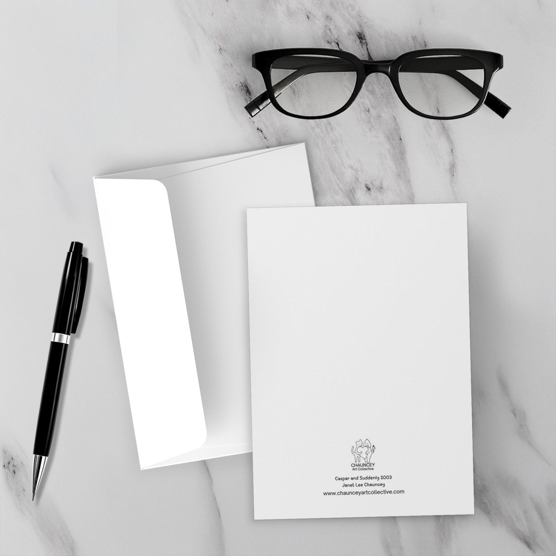 The backside of Caspar and Suddenly Greeting card with the title and Chauncey Art Collective logo. Displayed on a marble surface with a black pen and eyeglasses.