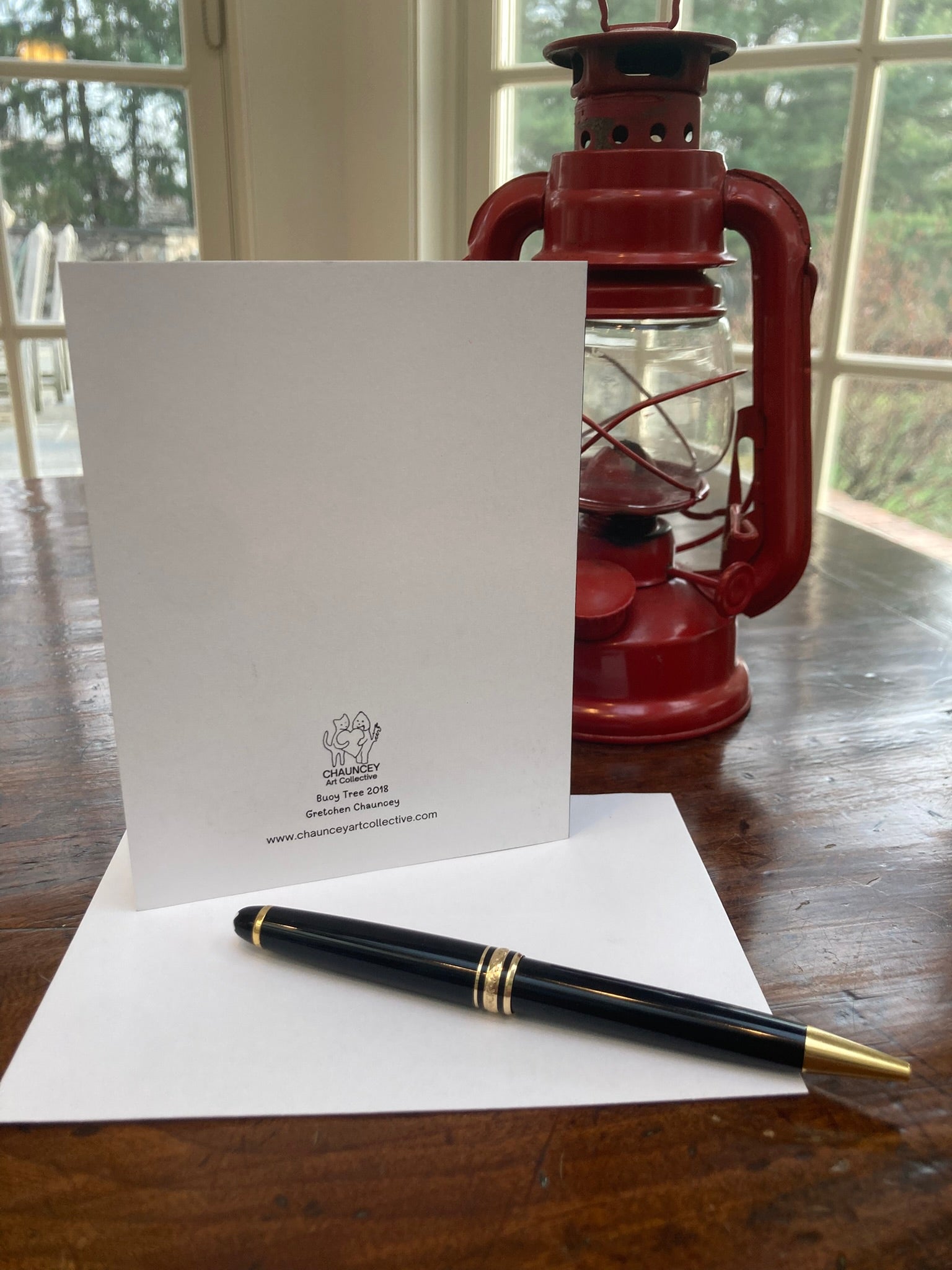 Backside of greeting card showing title and logo with a pen on a wooden surface with a red lantern in the background