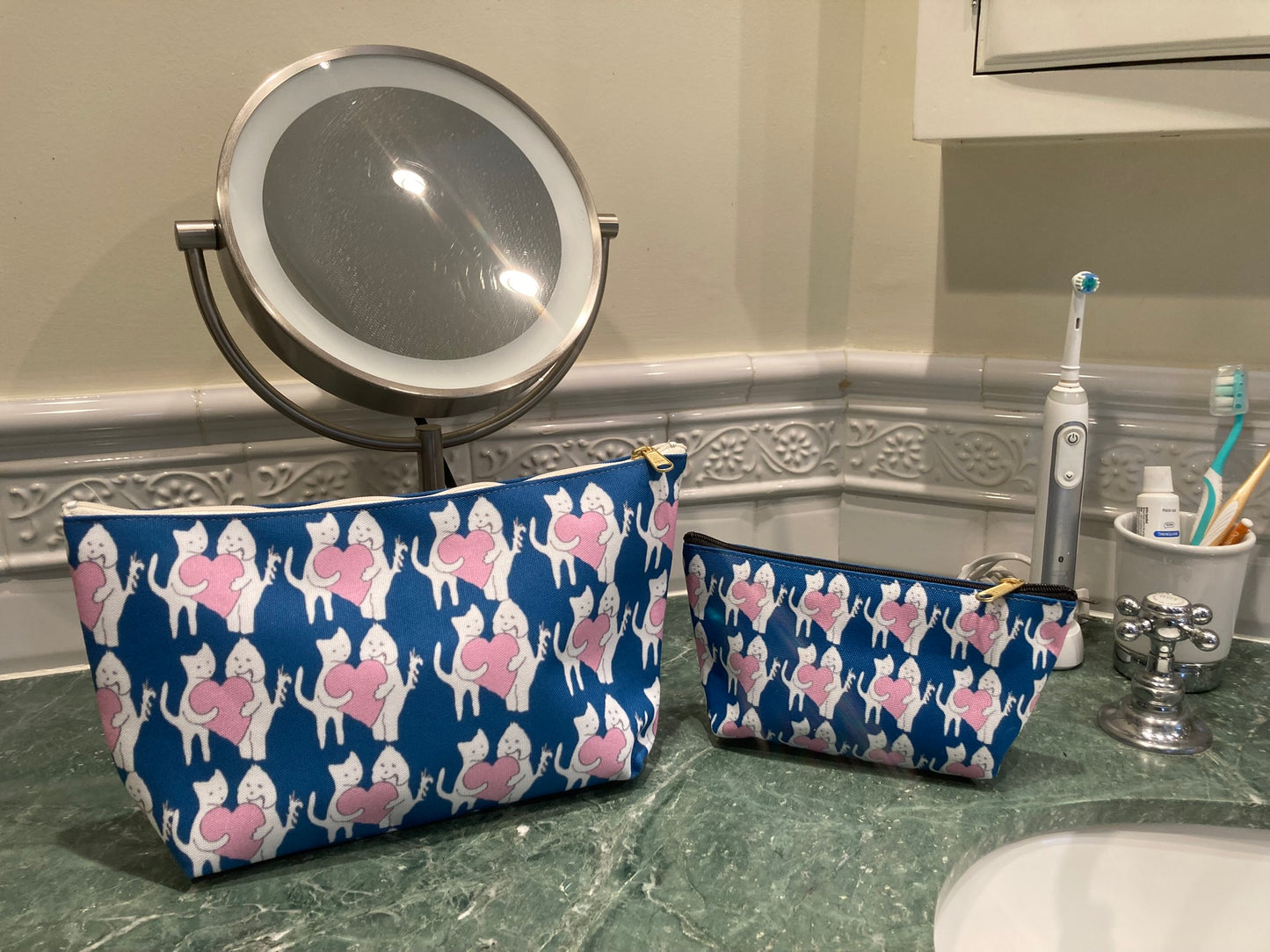 Two blue cosmetic bags with pink Chauncey Art Collective logo pattern on a bathroom counter.