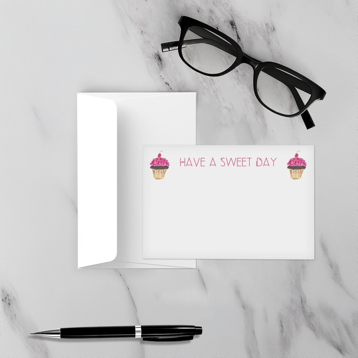 Note card with cupcake design and text Have a sweet day on a marble surface with glasses and pen.