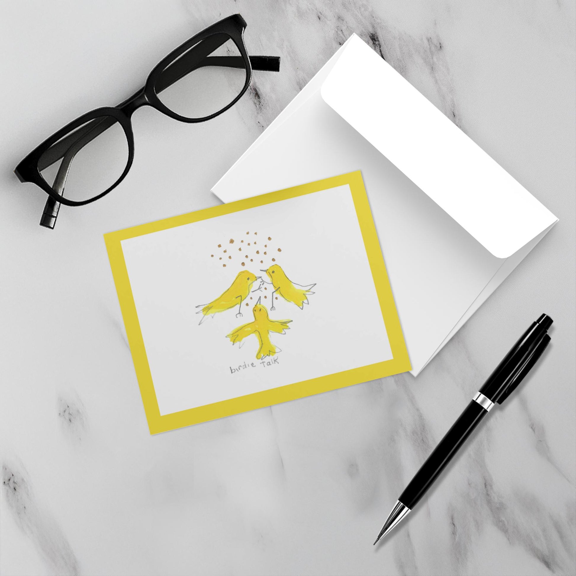 Mockup display showcasing the front of the greeting card Caspar Brings A Gift lying on a gray marble surface along side a white envelope, a pair of black reading glasses and a black pen.