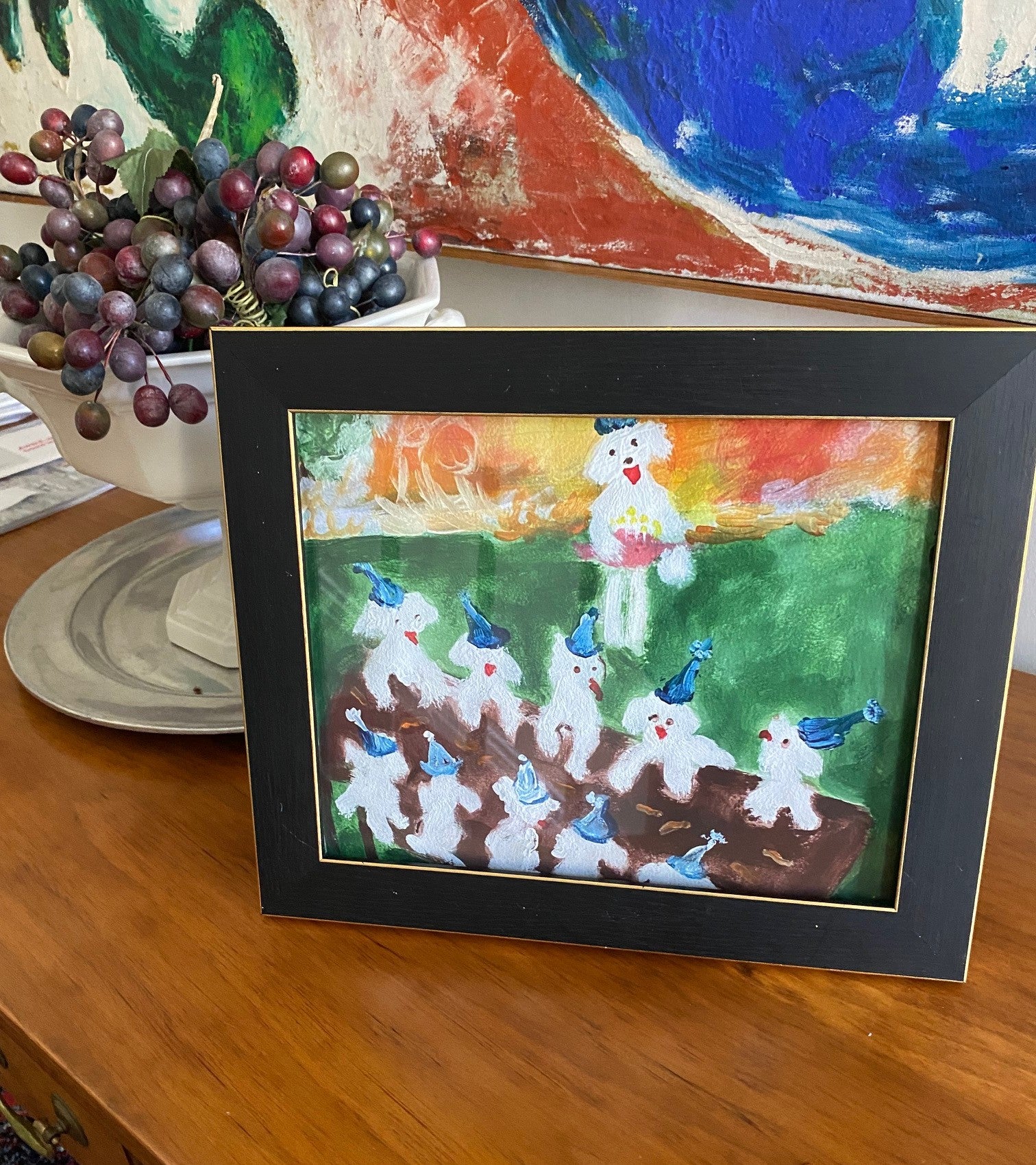 Mockup of Birthday Picnic art print in a black frame on a wood table next to a bowl of grapes. The colors in the print exude happiness as do the little Maltese doggie party goers.