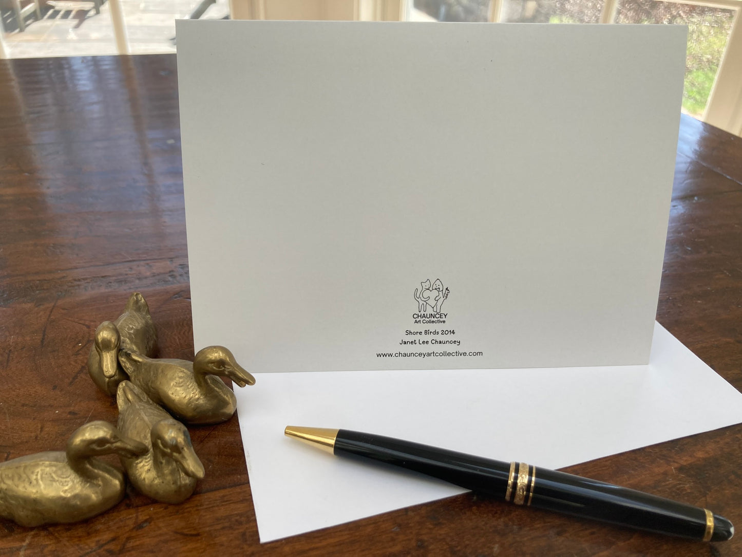 Reverse side of Shore Birds greeting card with logo and text on a wooden surface with gold duck decorations and a black pen.