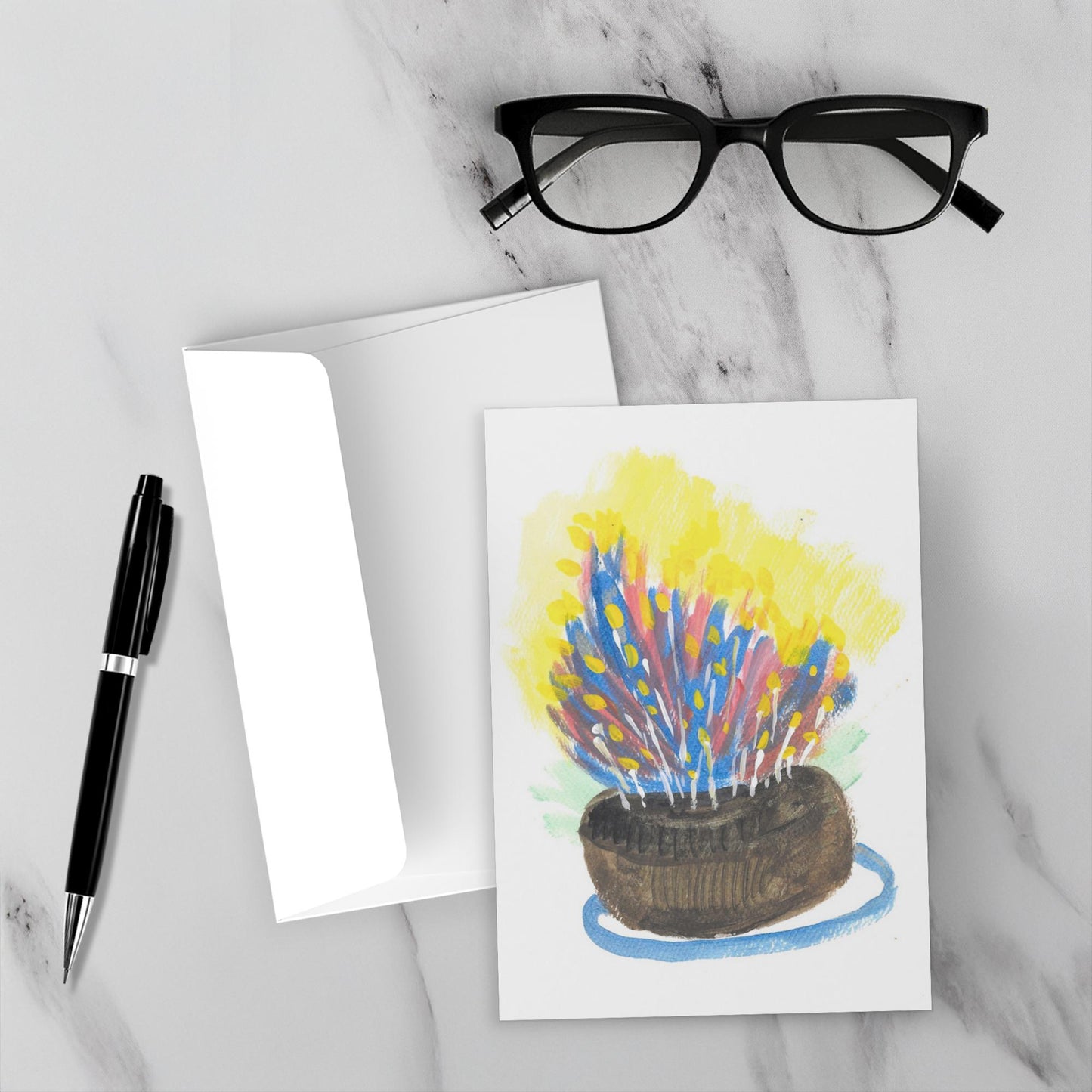 Mockup display showcasing the front of the greeting card The Most Delicious Cake lying on a gray marble surface along side a white envelope, a pair of black reading glasses and a black pen.