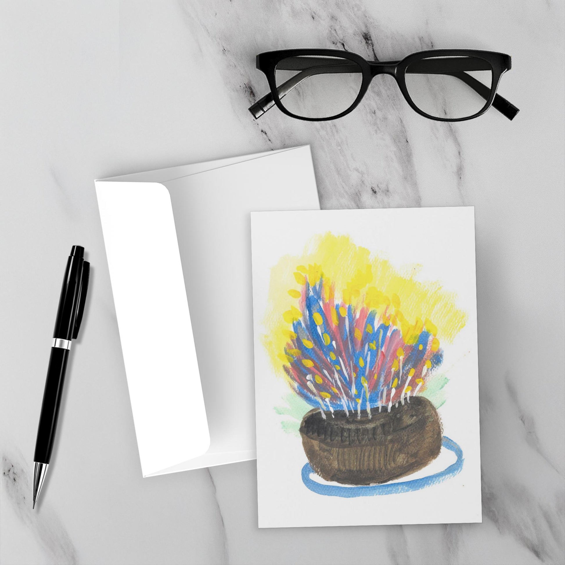 Mockup display showcasing the front of the greeting card The Most Delicious Cake lying on a gray marble surface along side a white envelope, a pair of black reading glasses and a black pen.