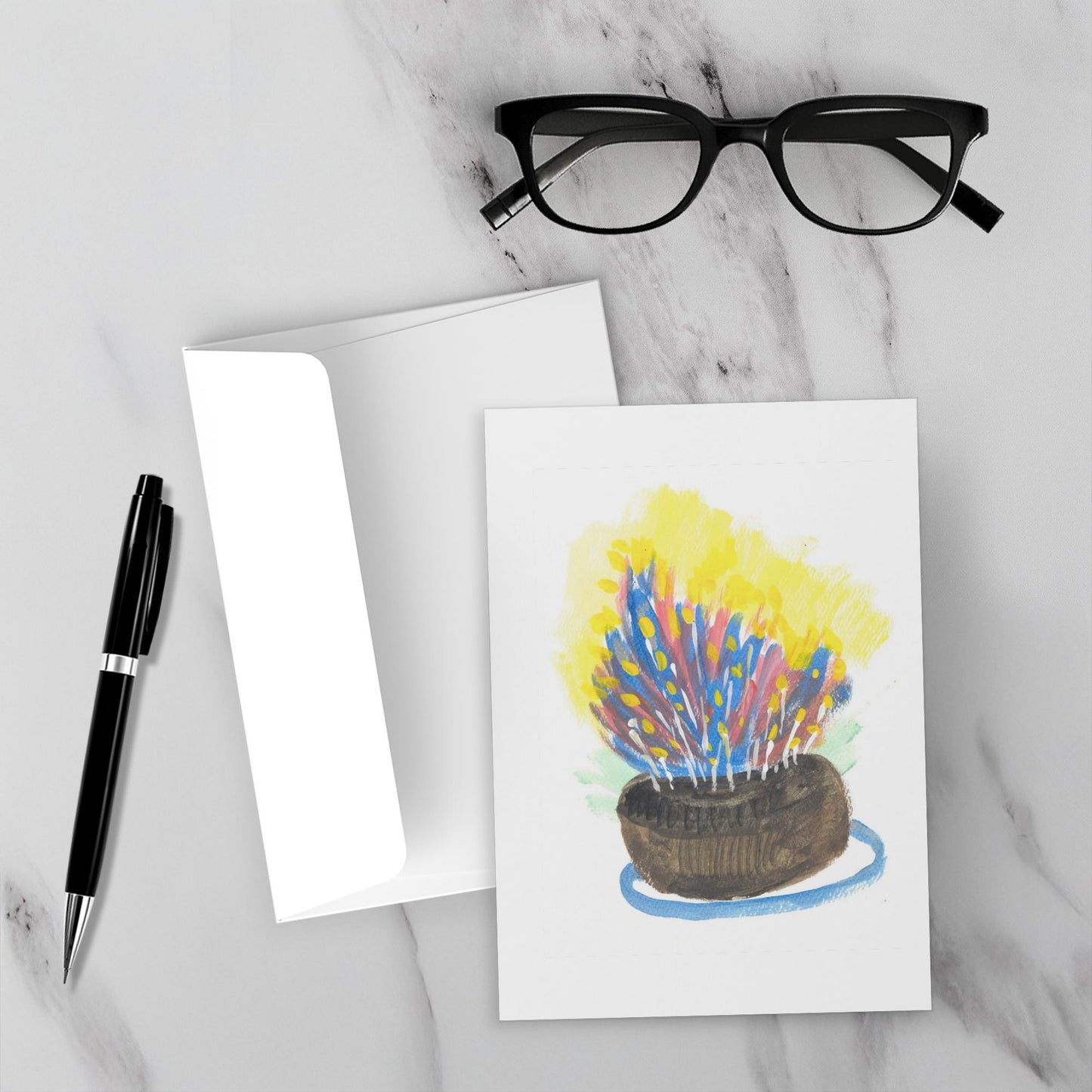 The Most Delicious Cake celebration card sits on a white marble surface with its envelope, a pen and eyewear.