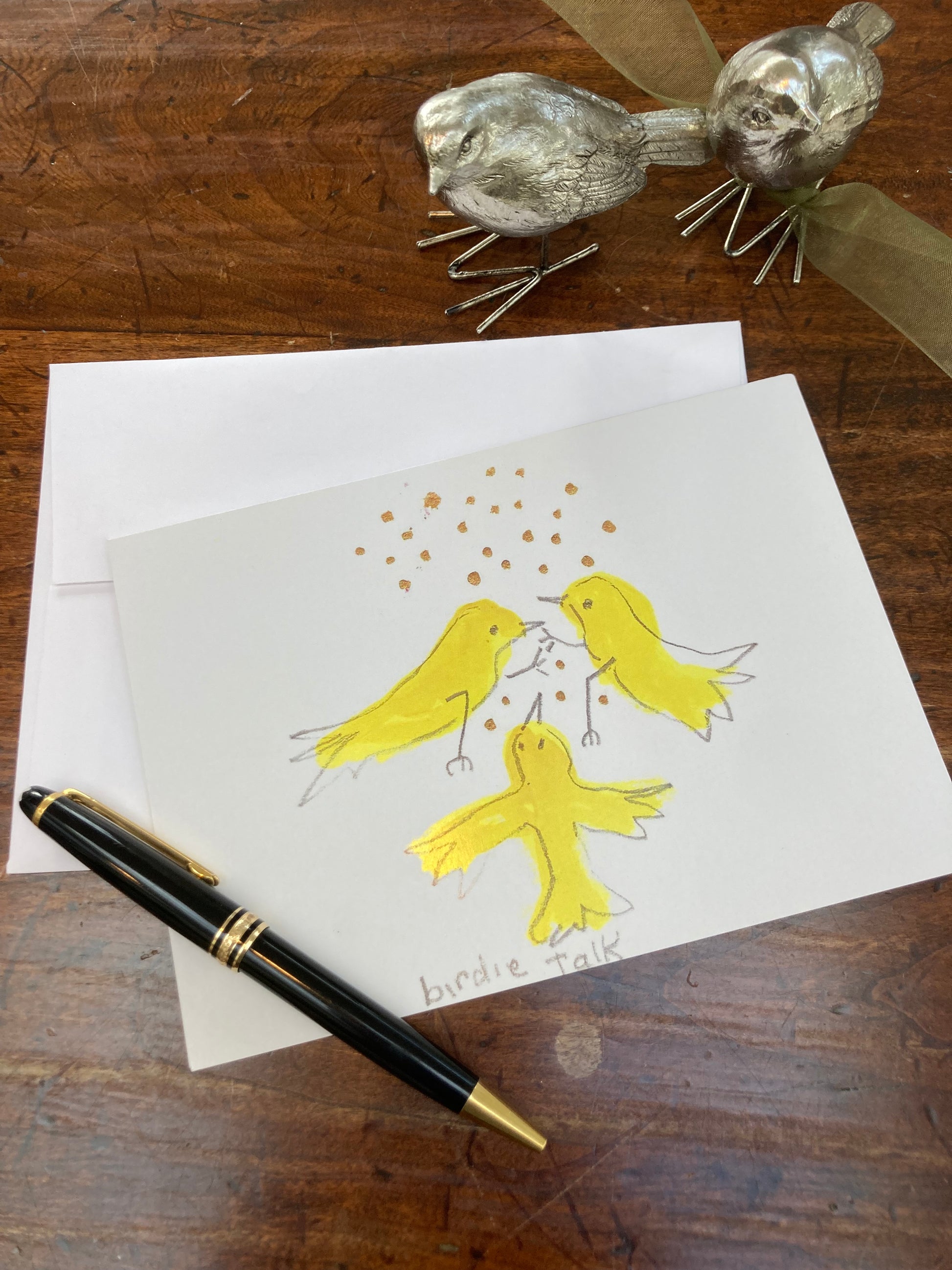Display of the front of Birdie Talk greeting card lying on the brown desk top with partial view of white envelope, a black fountain pen and two metal bird figurines.