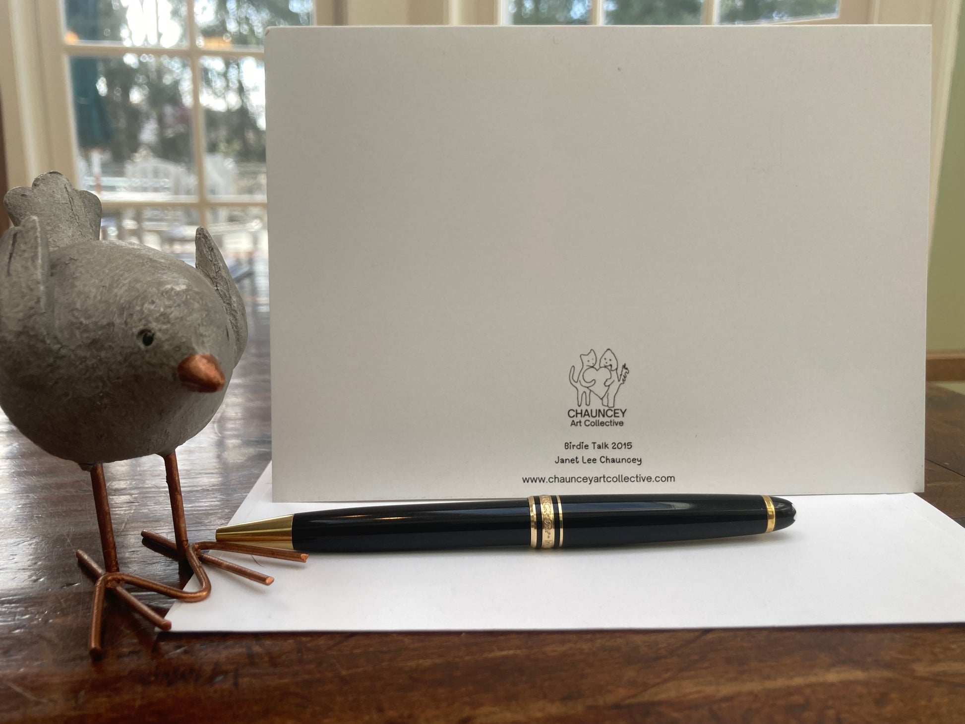 The back of the card Caspar Brings  Gift displayed standing upright on a brown desk with a white envelope and a black pen in front and a metal bird figurine to the left.