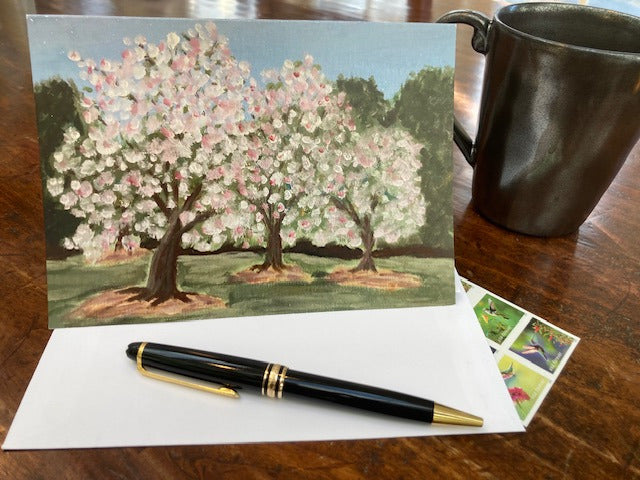 Mockup of the front of the greeting card situated on a wooden table with the envelope, stamps and a pen. A pewter coffee mug sits behind the card.