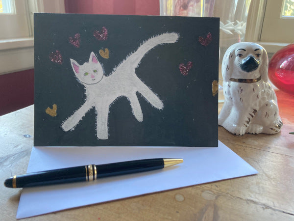 Front of the folded card Caspar With Floating Hearts is displayed on a brown desk with a white envelope, gold pen, and black and white Staffordshire dog statue to the right.